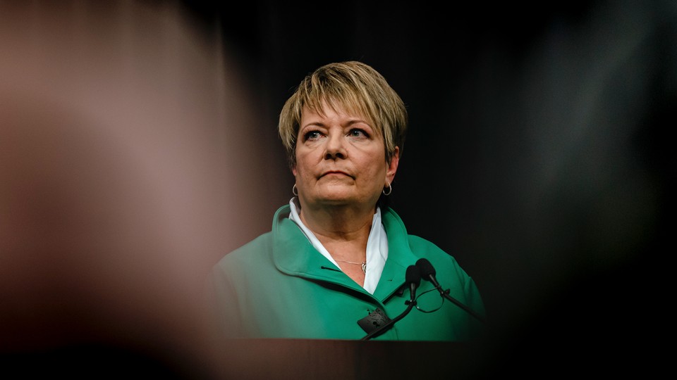 A photograph of Janet Protasiewicz, a short-haired blond woman, in a green jacket standing in front of a microphone